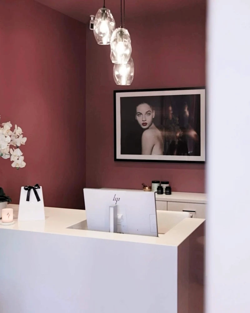 All Saint Clinic in Balmain in Australia reception scene with a white reception desk, a computer monitor, a framed black-and-white portrait on a dusty rose wall, and three hanging glass pendant lights. 