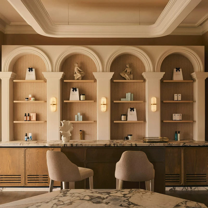All Saint Clinic in Mosman, Sydney Australia -  luxury spa / skin clinic interior featuring a marble counter, two upholstered bar stools, and built-in arched wooden shelves with decorative statues and skincare products. The color palette is dominated by warm beige, cream, and brown tones. 