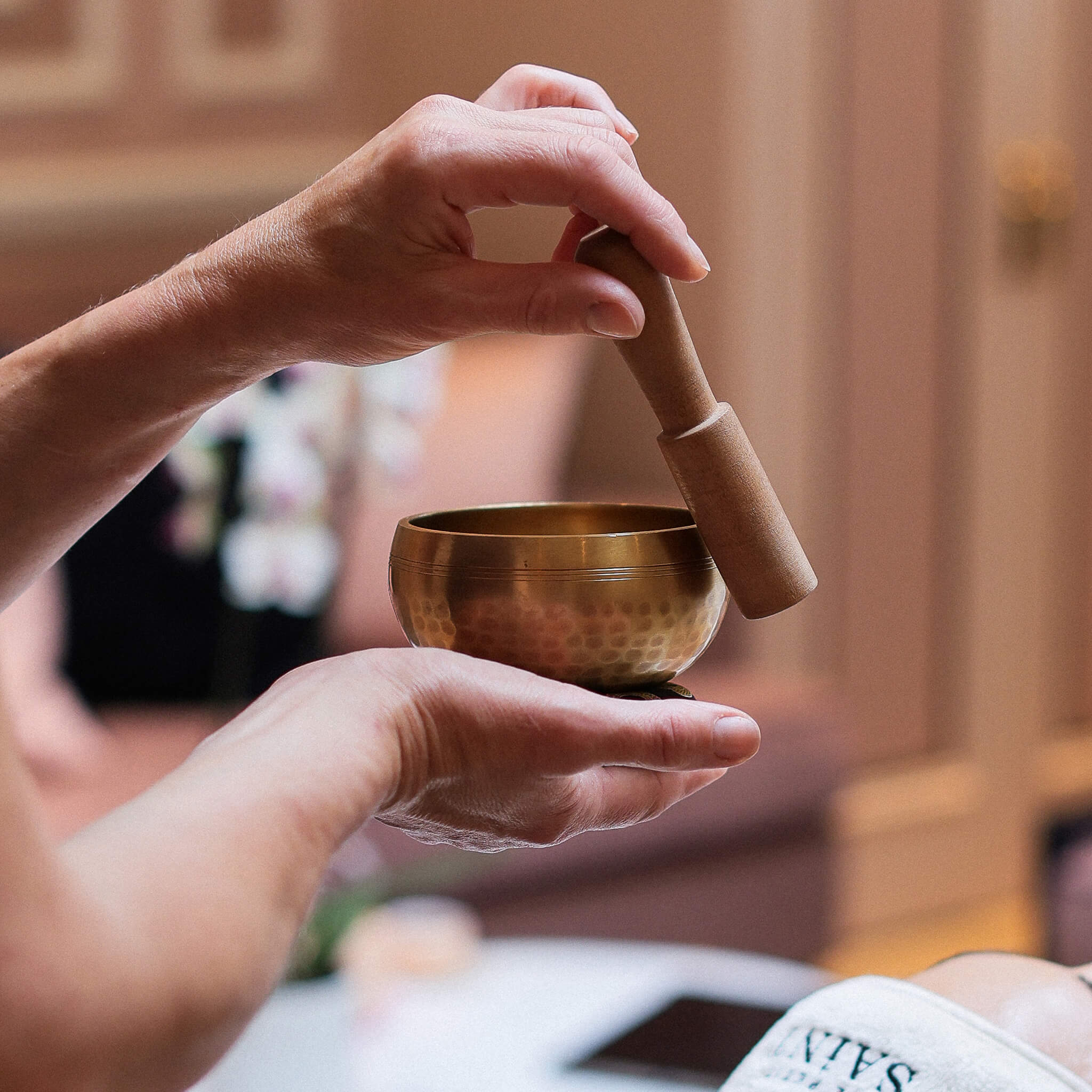 Person holding a bronze singing bowl with a wooden striker in a blurred indoor setting