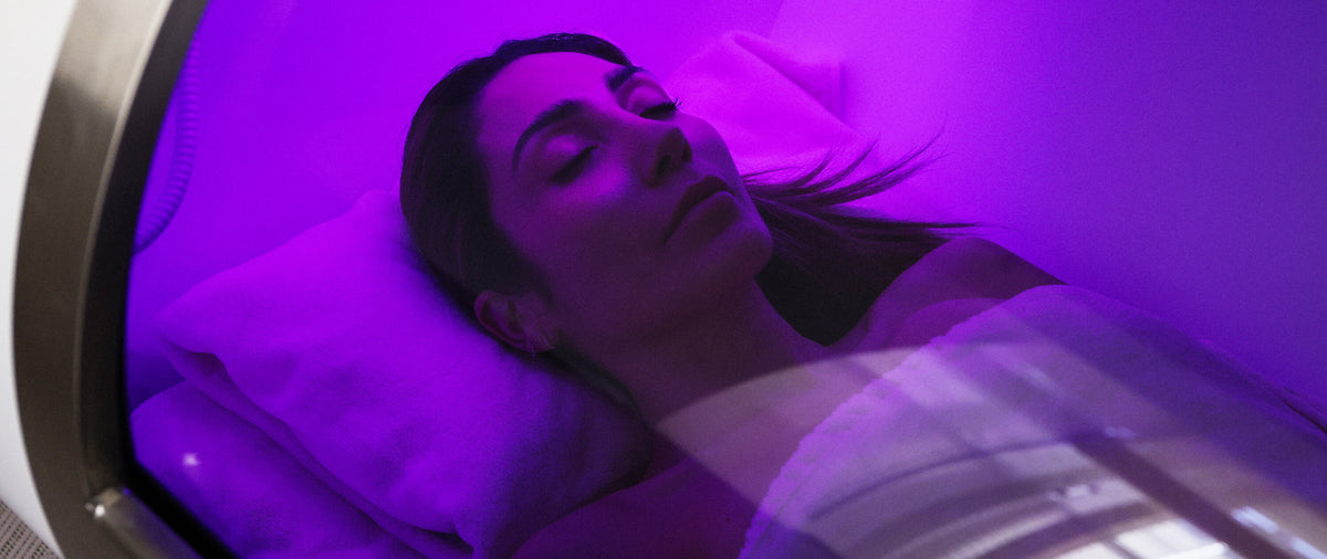 A woman lying down inside a Hyperbaric Oxygen Therapy bed under purple UV light, with pillows visible and the interior of the bed glowing magenta at Le Petit Saint Mayfair London.