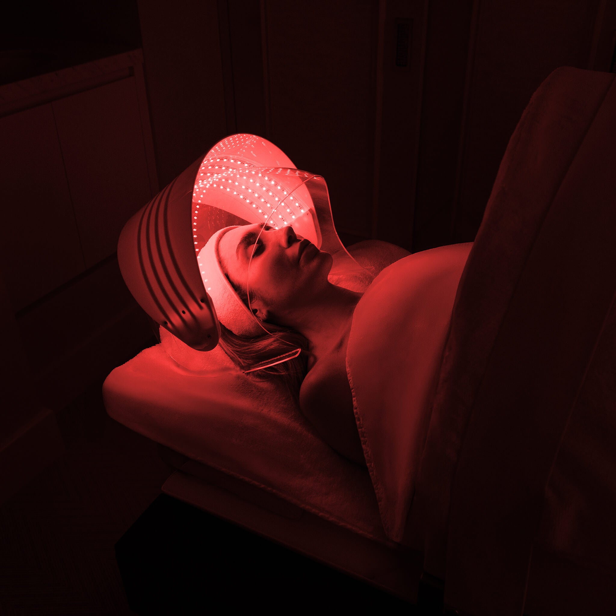 A woman lies on a treatment bed under a red LED light therapy helmet in a dim room, illuminated by the glowing red lights of the device.